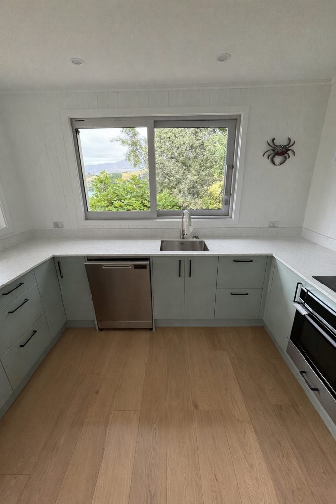 Modern white kitchen with green lower cabinets, wooden flooring, and large window providing natural light